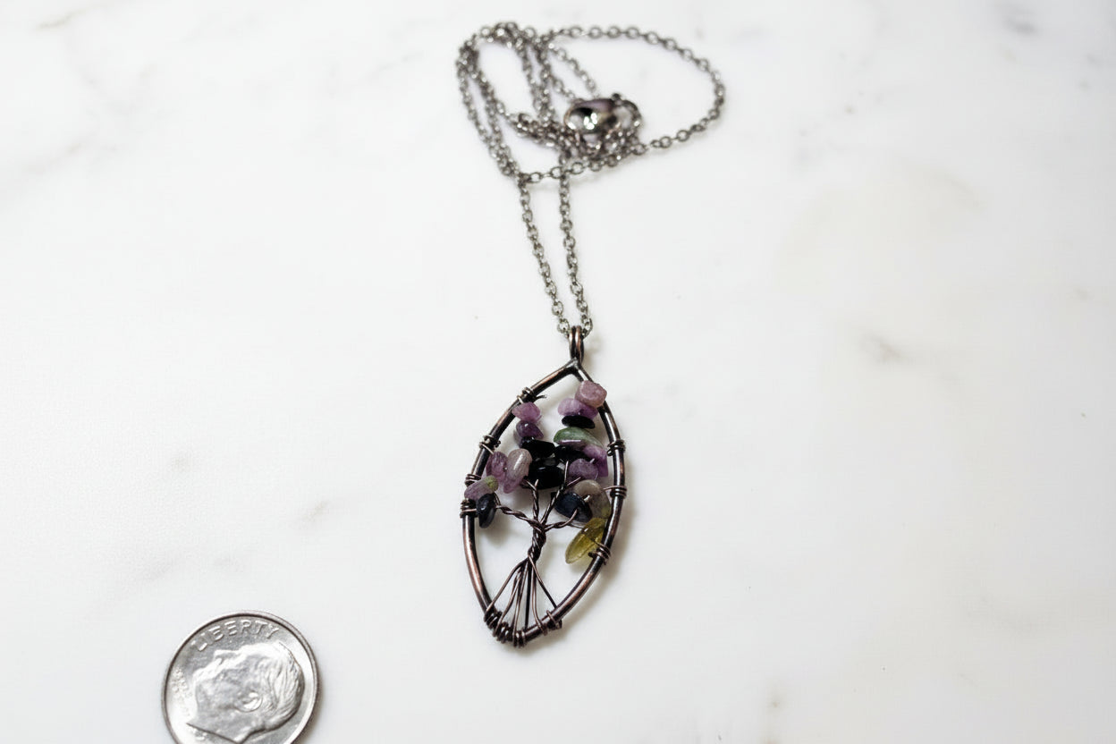 A silver-colored wire necklace with a tree of life pendant adorned with multicolored gemstones, displayed against a marble background with a coin beside it for scale.